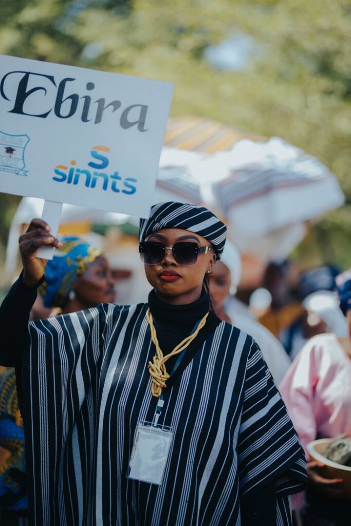 Woman in traditional African clothing holding sign reading 'Ebira' at vibrant outdoor event.